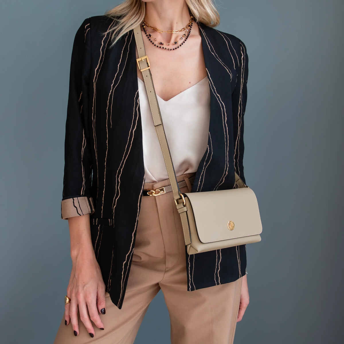 A woman models a light taupe crossbody handbag with gold hardware, worn over her shoulder. She is wearing a navy blue striped blazer, a cream camisole, and tan trousers against a solid slate gray background.