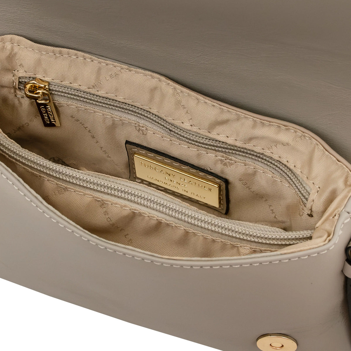 Close-up of light taupe handbag interior showing a zippered pocket with a gold-tone metal zipper pull, gold-tone brand plaque and beige lining.