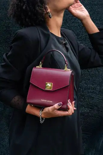 A woman in a black outfit holds a structured bordeaux leather handbag with gold hardware and a top handle against a dark textured background.