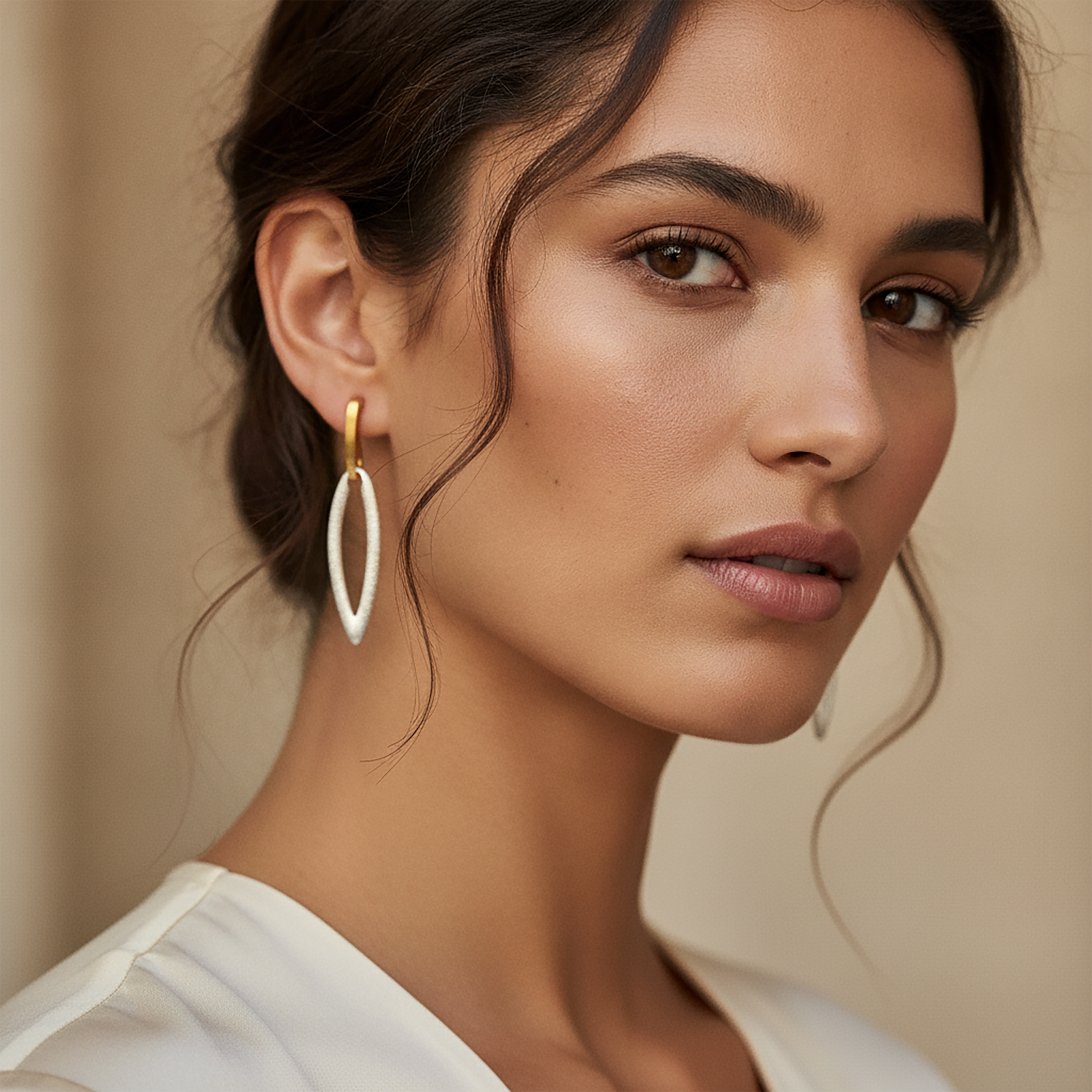 A close-up of a woman wearing a pair of large open oval drop earrings, one in polished gold and one in white enamel, set against a soft beige background.