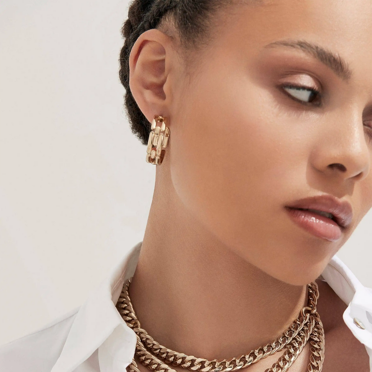 A close-up of a model wearing a pair of gold hoop earrings with chain interlock design, styled with layered gold chain necklaces and a white collared shirt.