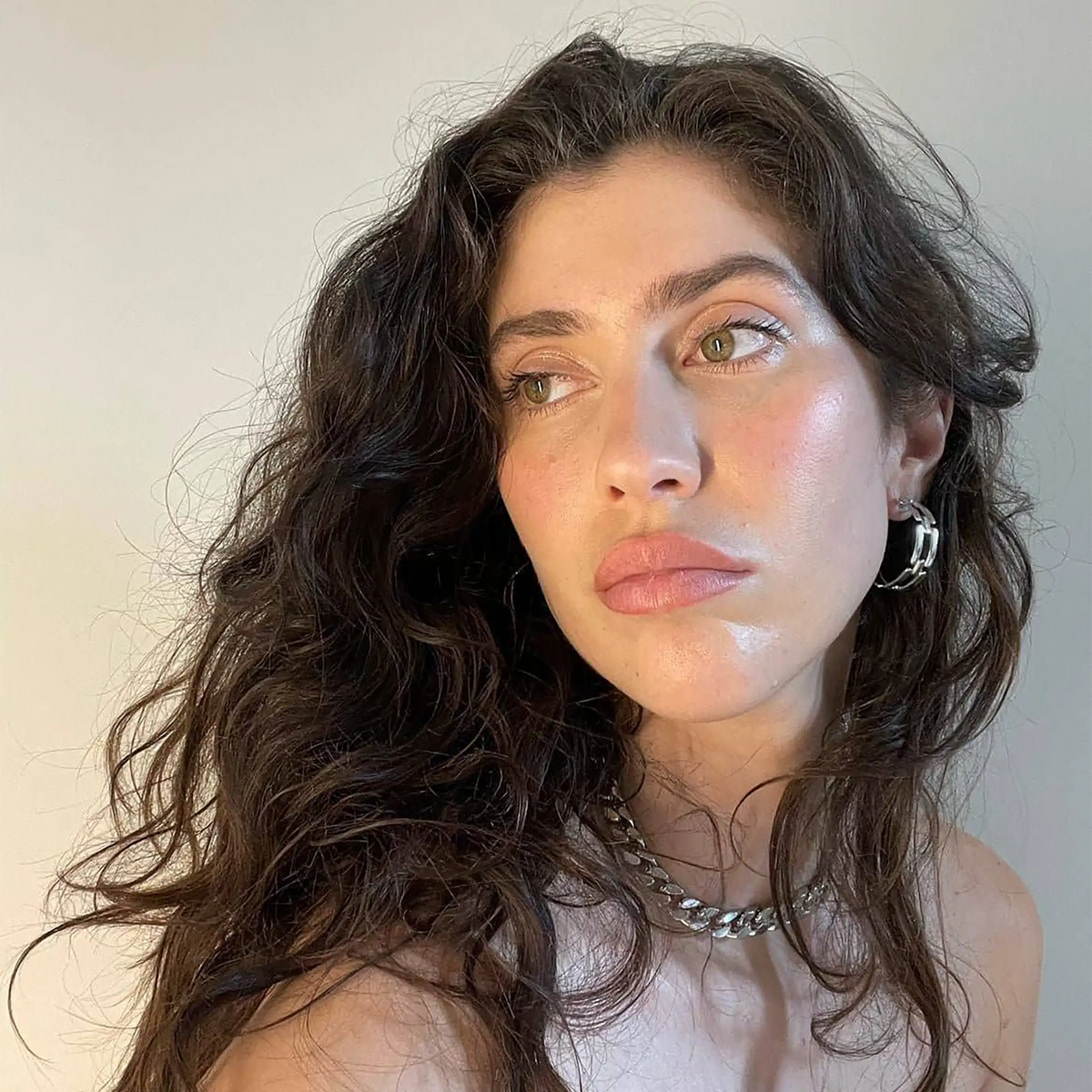 A close-up portrait of a woman with long, wavy dark brown hair, wearing a silver chain necklace and a silver chain interlock hoop earrings, set against a plain light background.