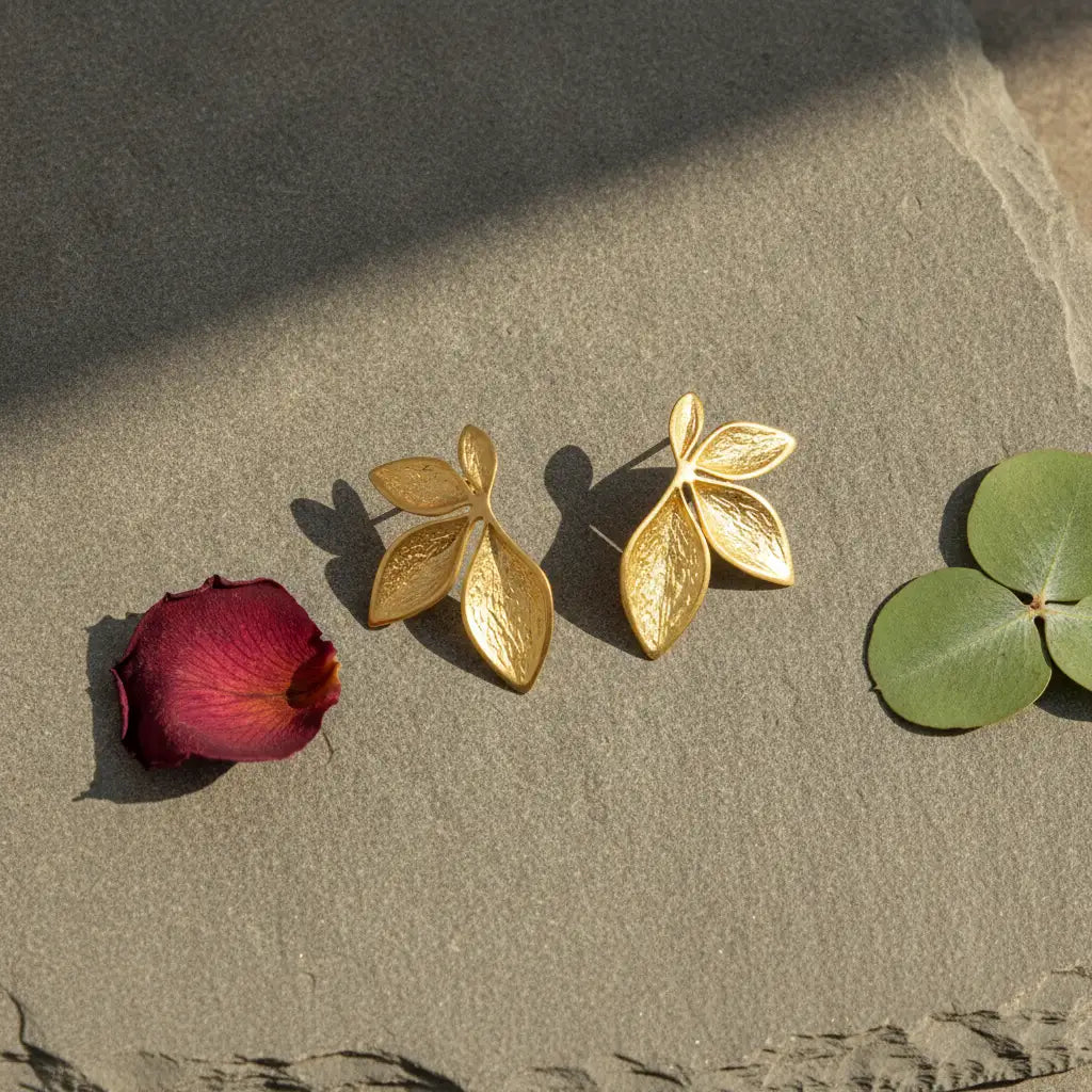 Two gold leaf-shaped stud earrings displayed on a textured stone surface, flanked by a red rose petal on the left and green leaves on the right.