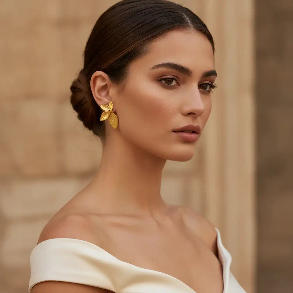A woman with her hair in a low bun wears a pair of gold leaf-shaped stud earrings, photographed in a warm, softly lit setting with a blurred stone background.
