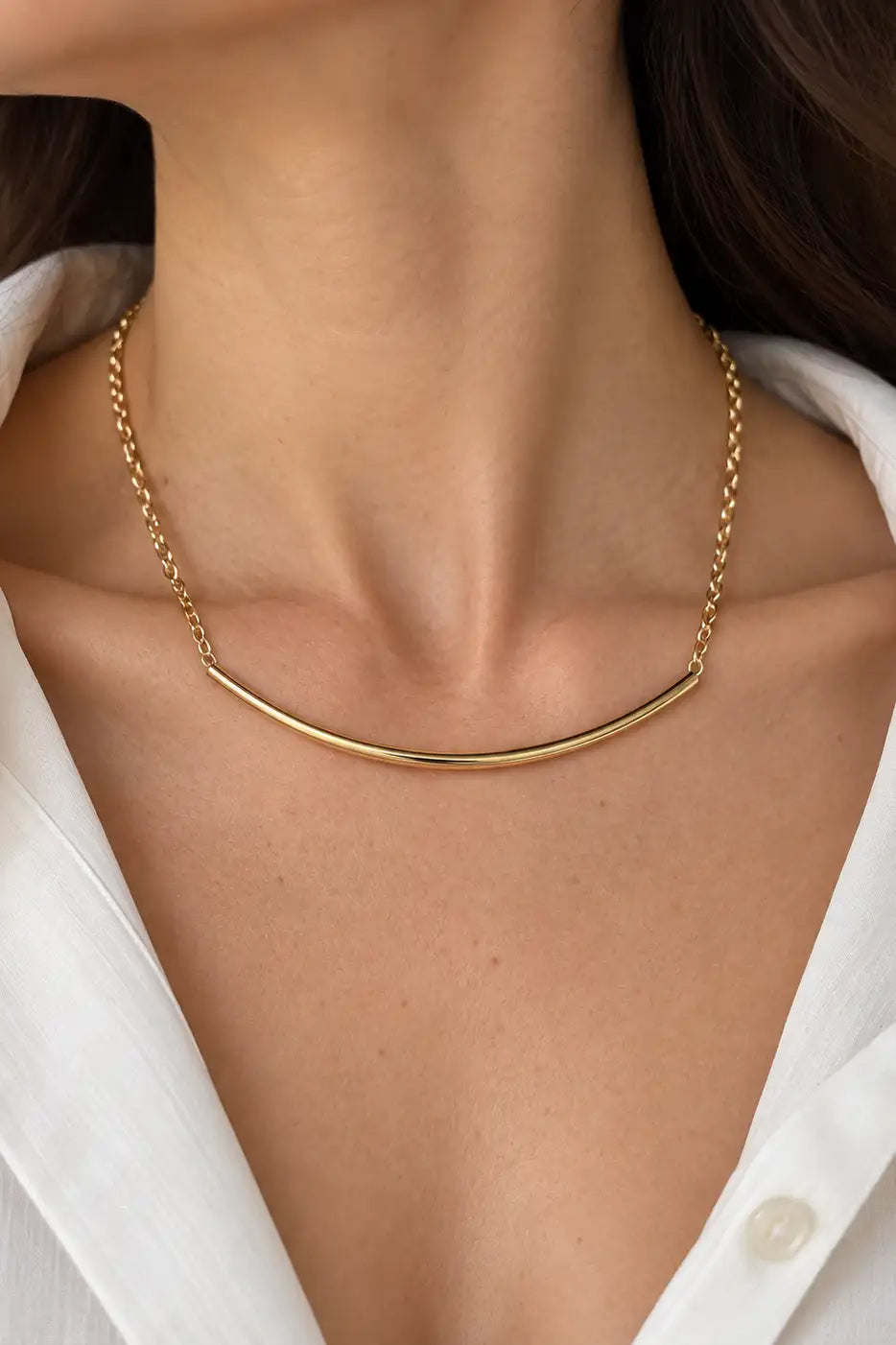 A close-up of a model's neck and upper chest wearing a delicate gold necklace with a curved bar design, paired with a white collared shirt.