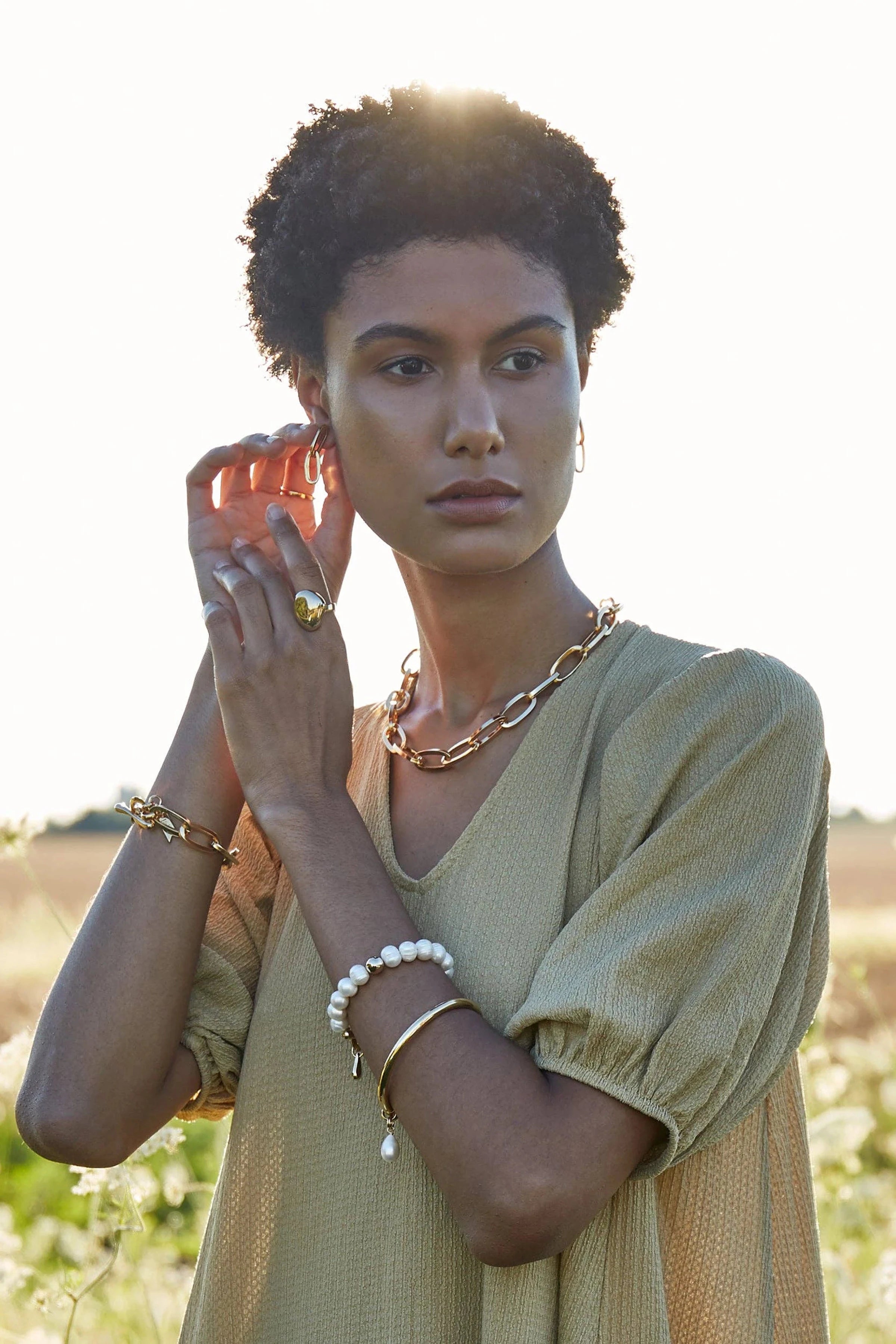 A woman wearing an olive v-neck blouse , golden chainlink jewelry on her neck, bracelet and fingers.