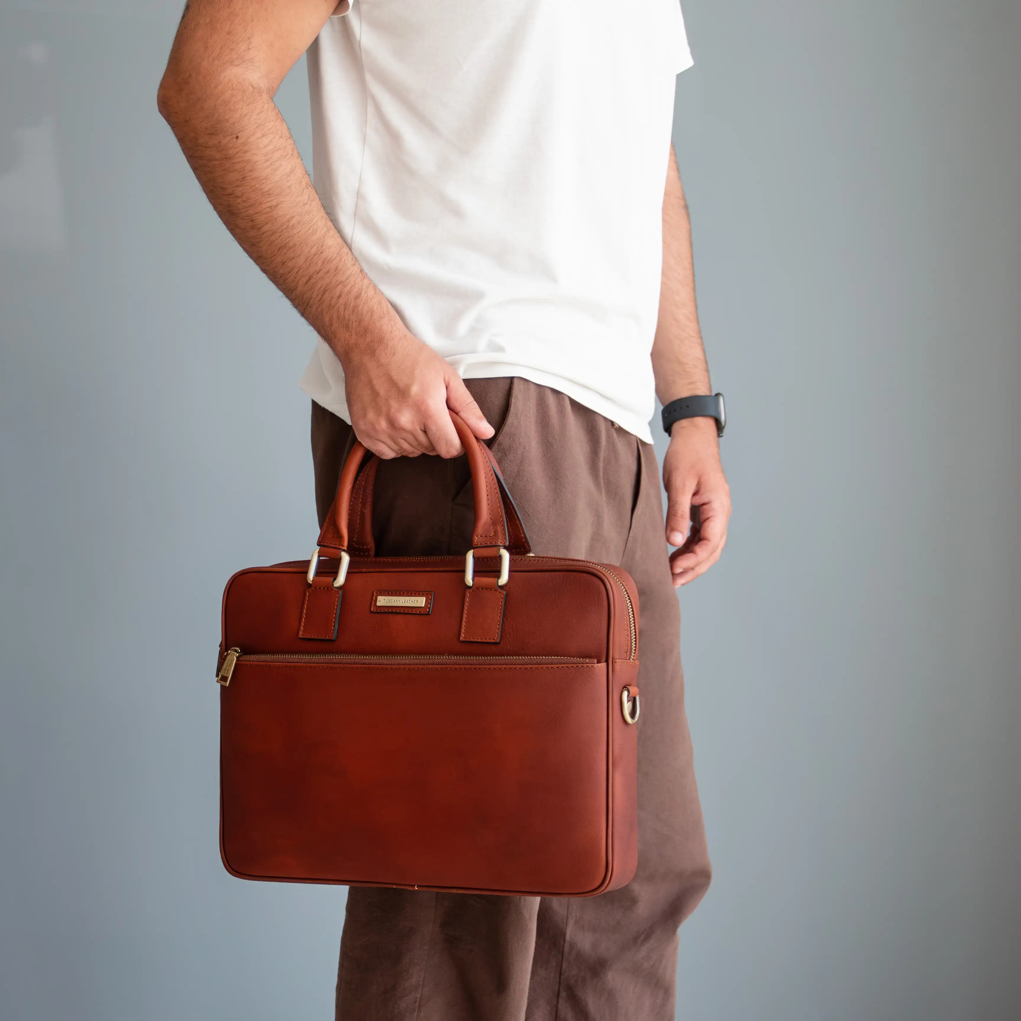 A man in a white t-shirt and brown trousers holds a brown leather briefcase with gold-tone hardware against a plain gray background.