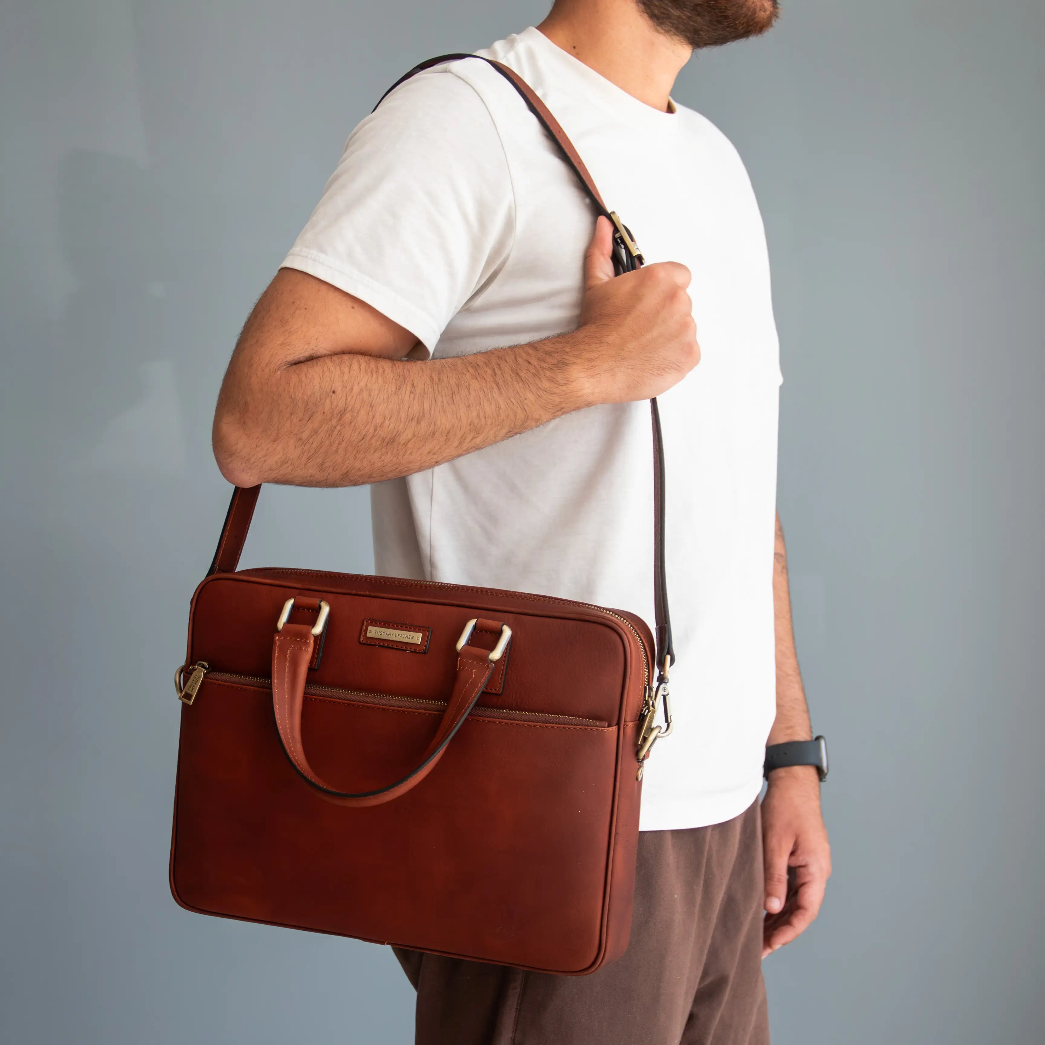 A man wearing a white t-shirt and brown trousers carries a reddish-brown leather briefcase-style shoulder bag with gold-tone hardware against a plain gray studio background.
