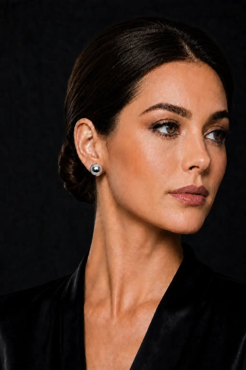 A close-up portrait of a woman with dark hair styled in an updo, wearing a single round silver-toned stud earring against a dark background.