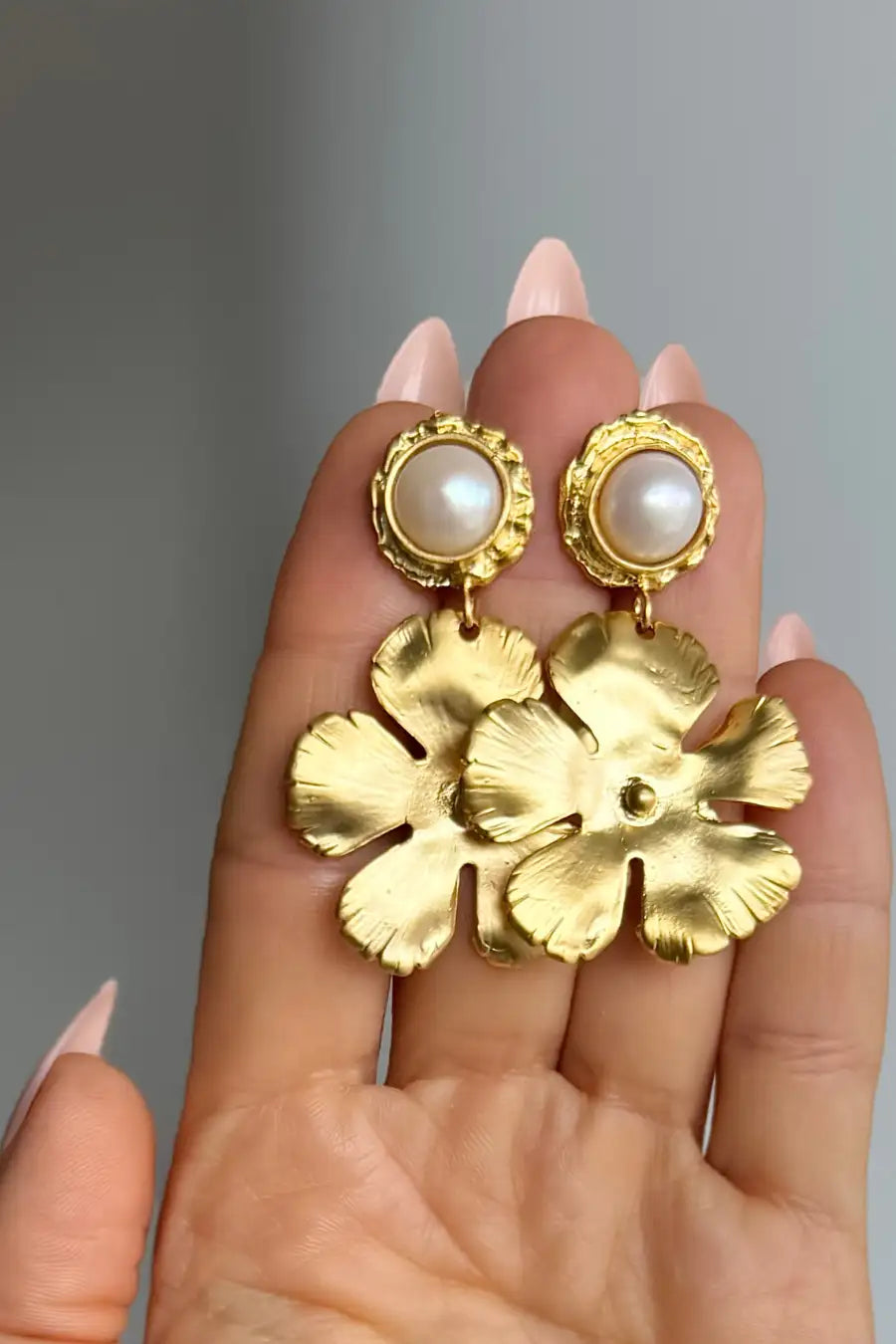 A pair of gold dangle earrings with pearl-like round studs and textured flower-shaped drops, held in a person's hand against a soft gray background.