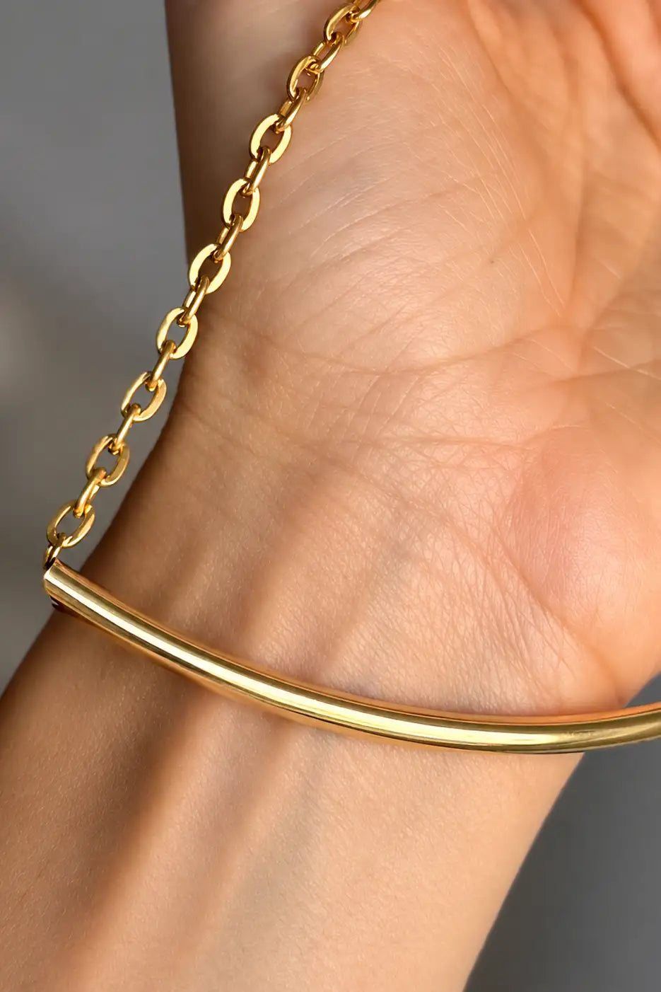 A close-up of a gold bangle bracelet worn on a wrist, featuring a polished curved bar linked to a delicate gold chain.