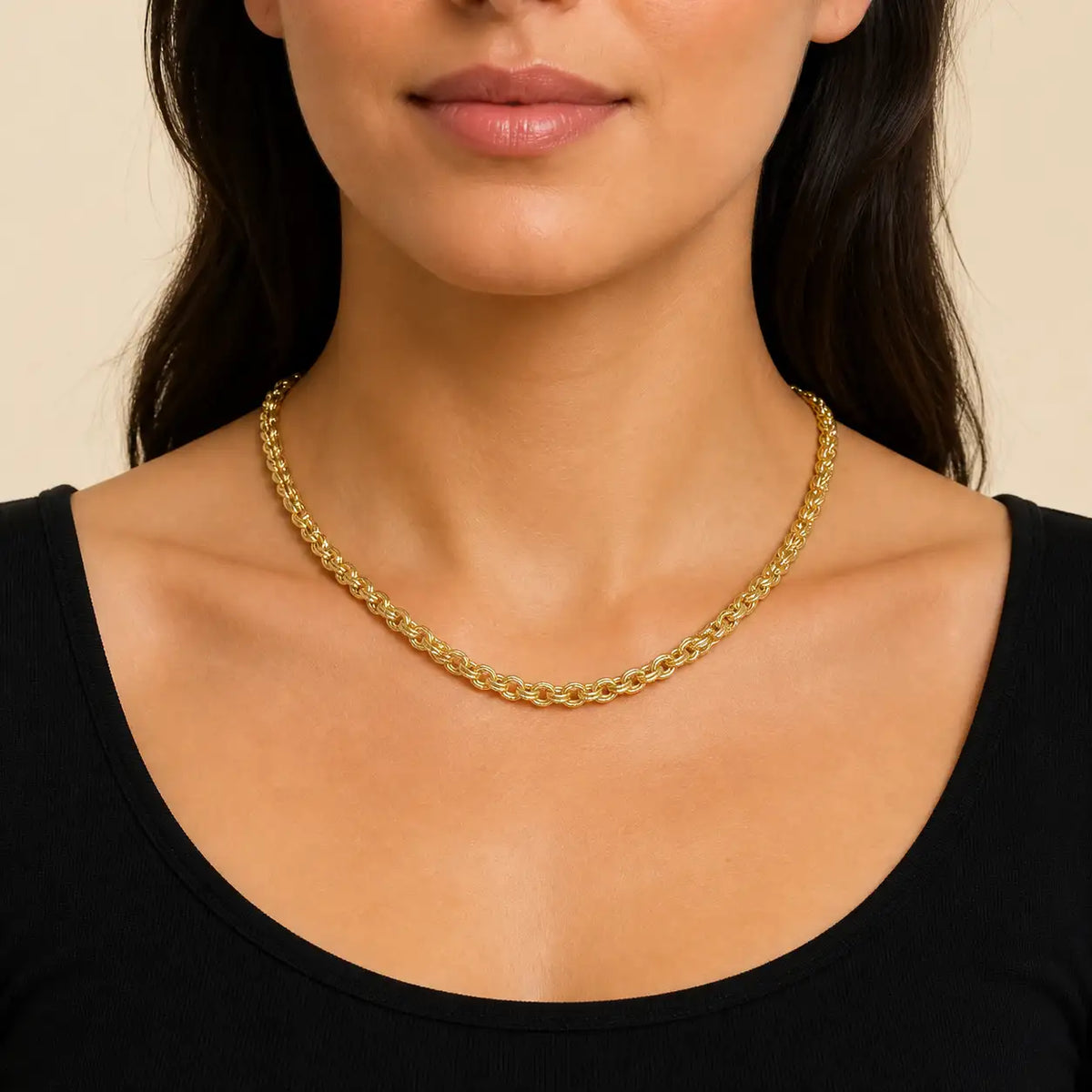 A close-up of a model wearing a gold chain necklace with a thick, textured curb link design, paired with a black scoop-neck top against a warm beige background.