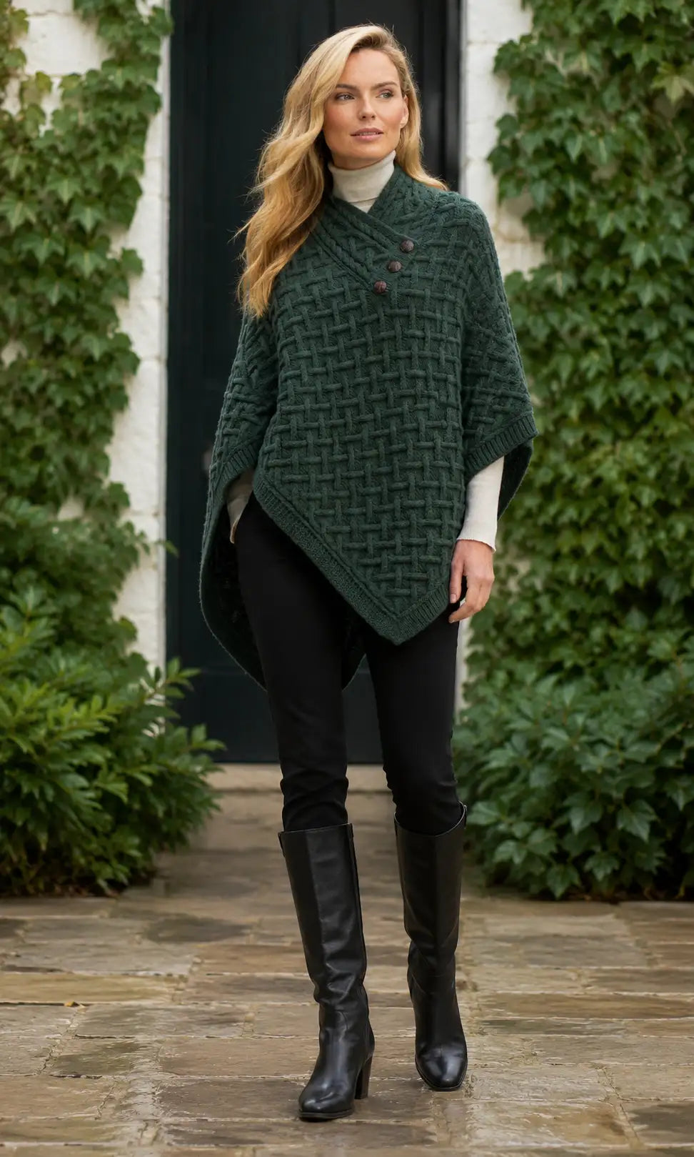 A woman wearing a dark green textured knit poncho over a white turtleneck, black pants, and black knee-high boots, standing on a stone path in front of a doorway with ivy-covered walls.