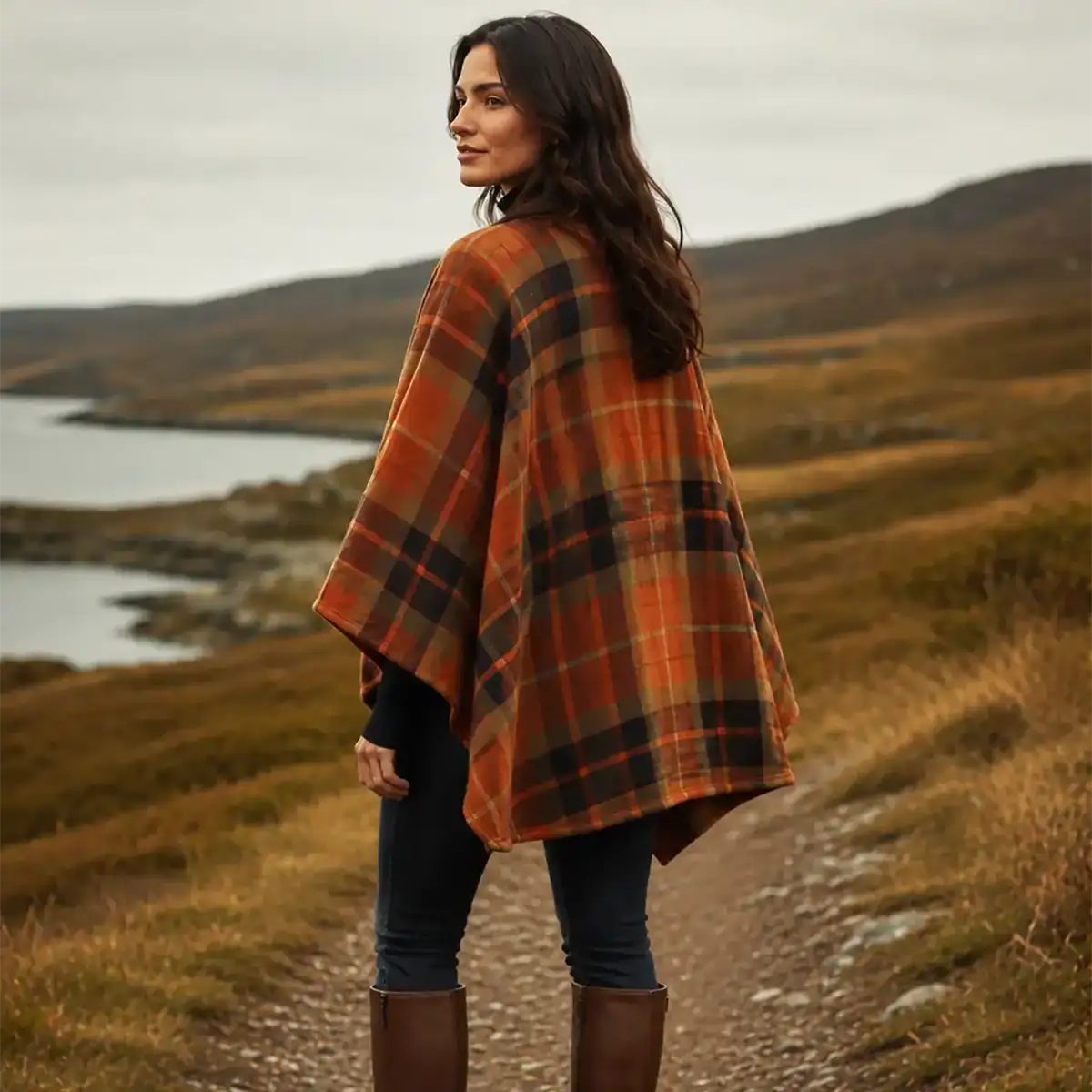 A woman stands on a dirt path in a rural landscape, wearing a rust-orange plaid poncho over dark jeans and tall brown boots.