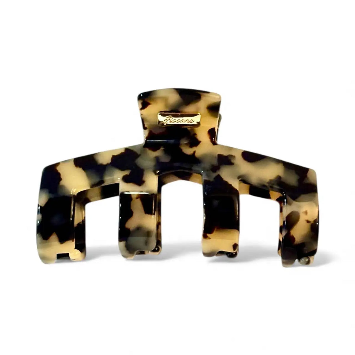 A large claw-style hair clip in a glossy tortoiseshell pattern of black, brown, and beige, featuring a small gold-tone metal brand plaque on the top bar.