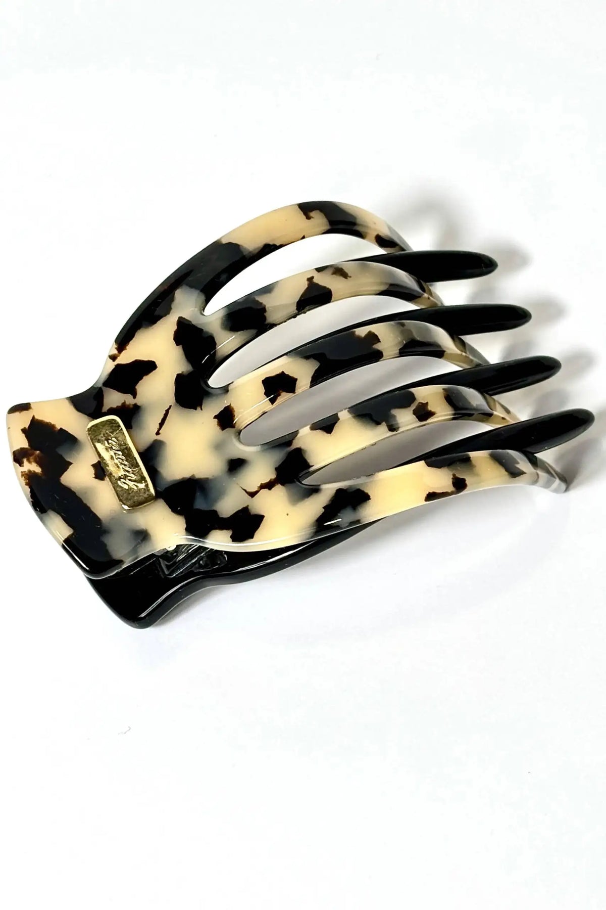 A tortoiseshell claw hair clip with a cream and black mottled pattern, featuring a small gold-tone brand plaque on the side, set against a plain white background.