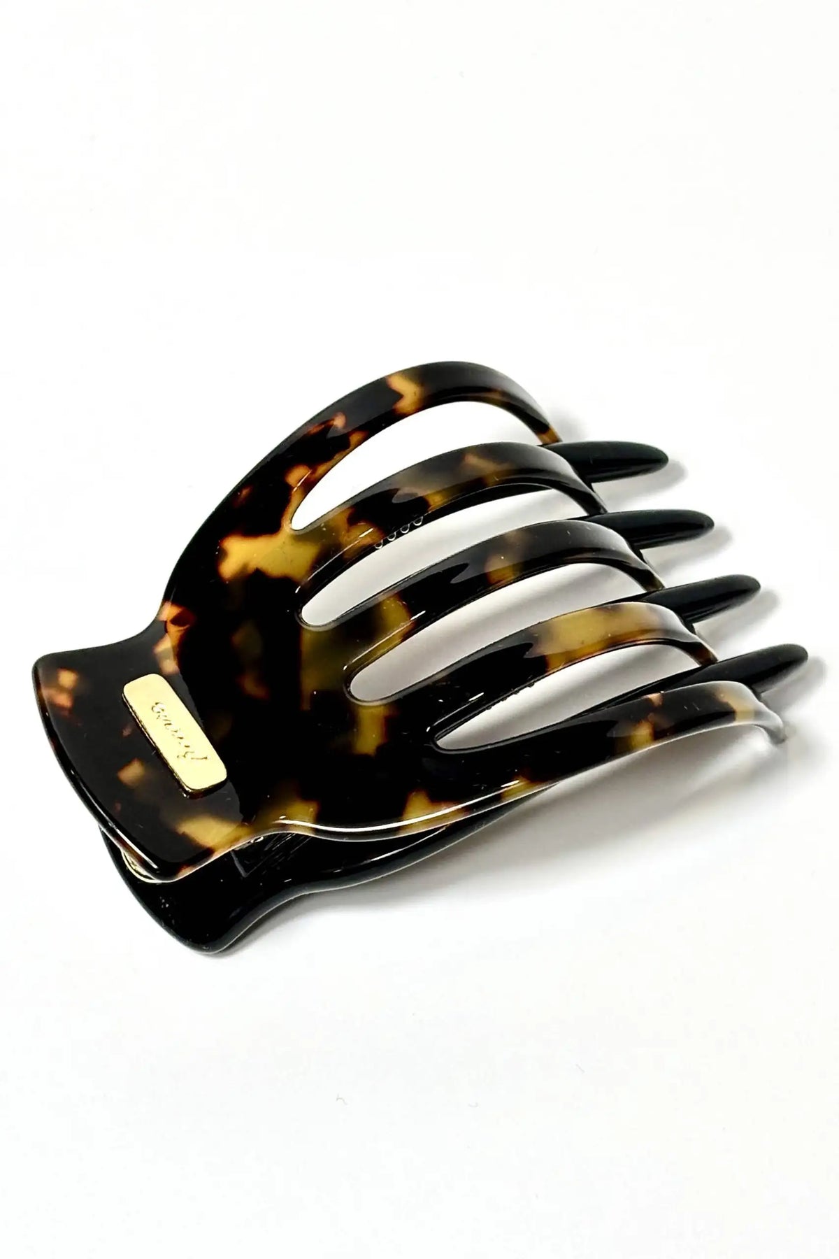 A large hair claw clip in a glossy dark brown and amber-brown tortoiseshell pattern, featuring a small gold-tone metal brand plaque that reads 'LUXURY'.