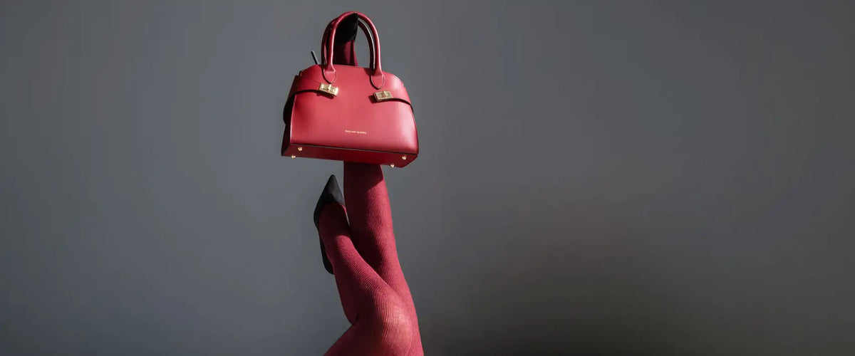Red Tuscany Leather handbag with gold-tone hardware balanced on raised legs wearing burgundy tights and black heels against a dramatic gray background.