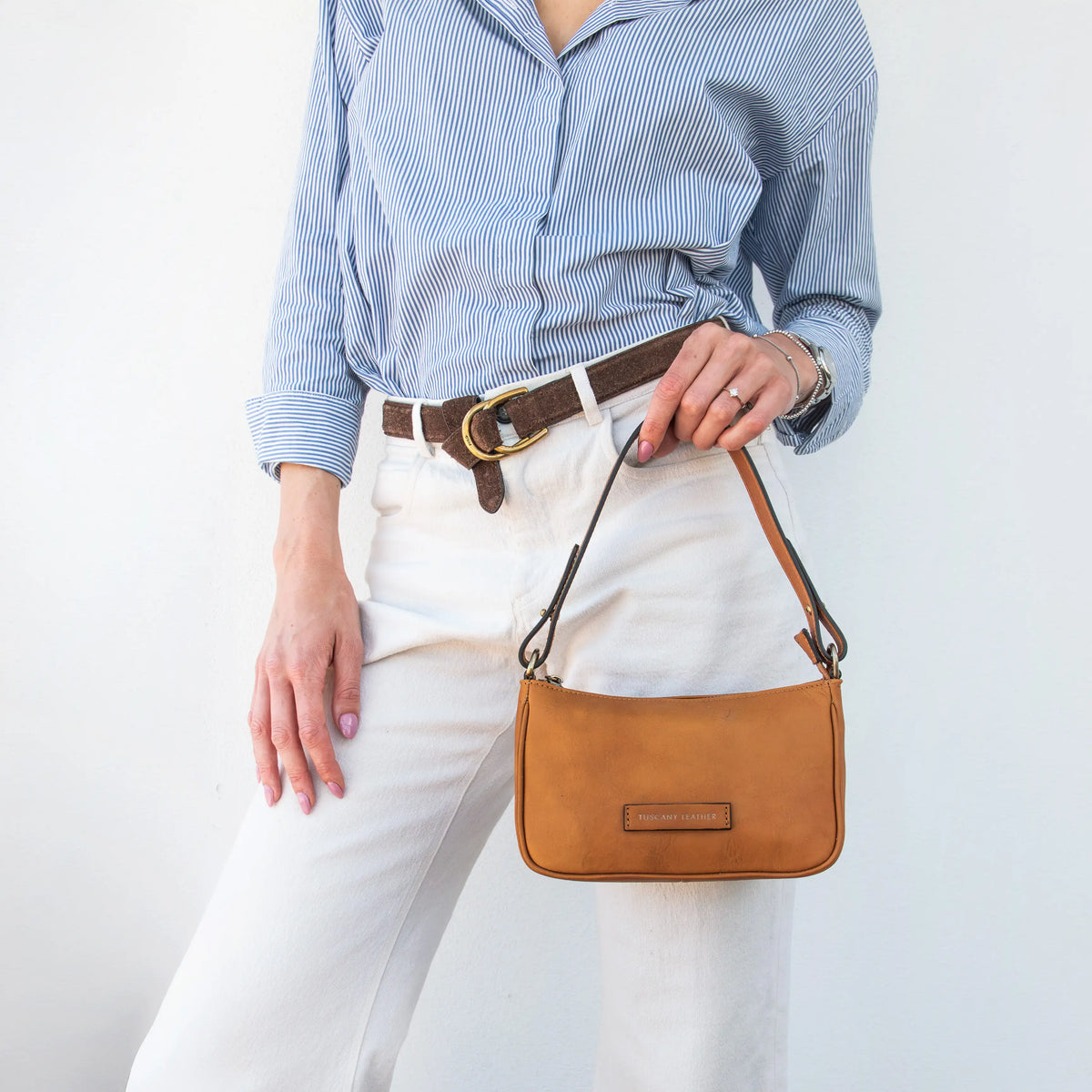A person wearing a blue and white striped button-down shirt, white pants, and a brown belt holds a small natural leather shoulder bag with a brown strap and gold-tone buckle against a plain white background.