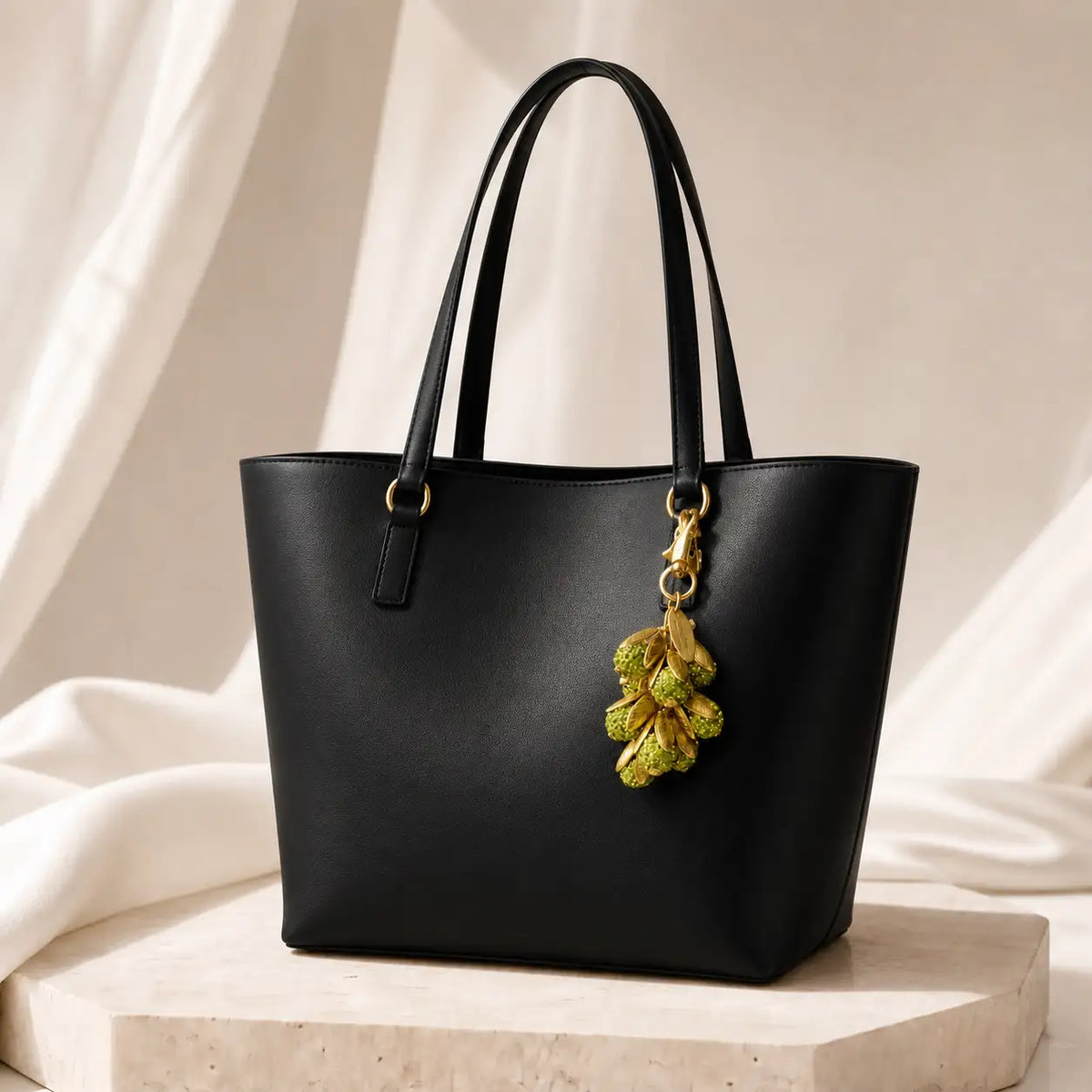 A black leather tote bag with double shoulder straps and a gold-tone hanging ornament featuring green leaves, displayed on a stone pedestal against a draped cream fabric background.