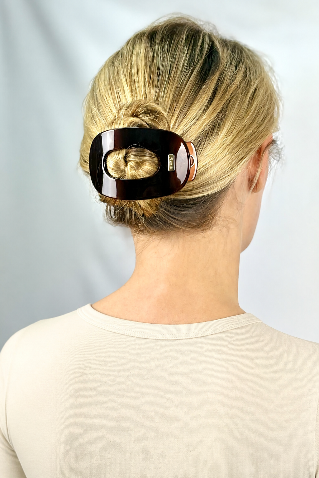 A woman viewed from the back wearing a large, glossy brown flat hair claw clip holding her blonde hair in a low bun. 