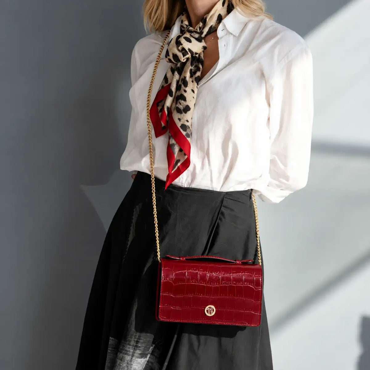 A woman wearing a white blouse, black skirt and leopard-print scarf carries a red crocodile-embossed shoulder bag with a gold chain strap as a crossbody.
