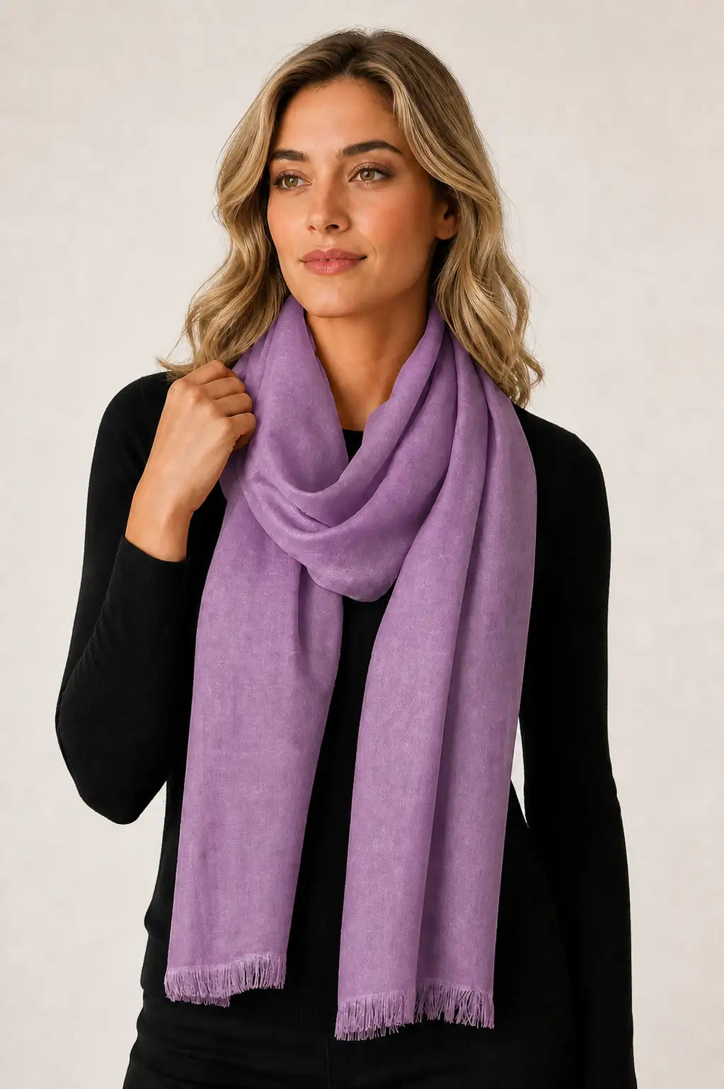 A woman with blonde hair wearing a soft lavender-purple scarf and a black long-sleeve top, posing against a light beige studio background.