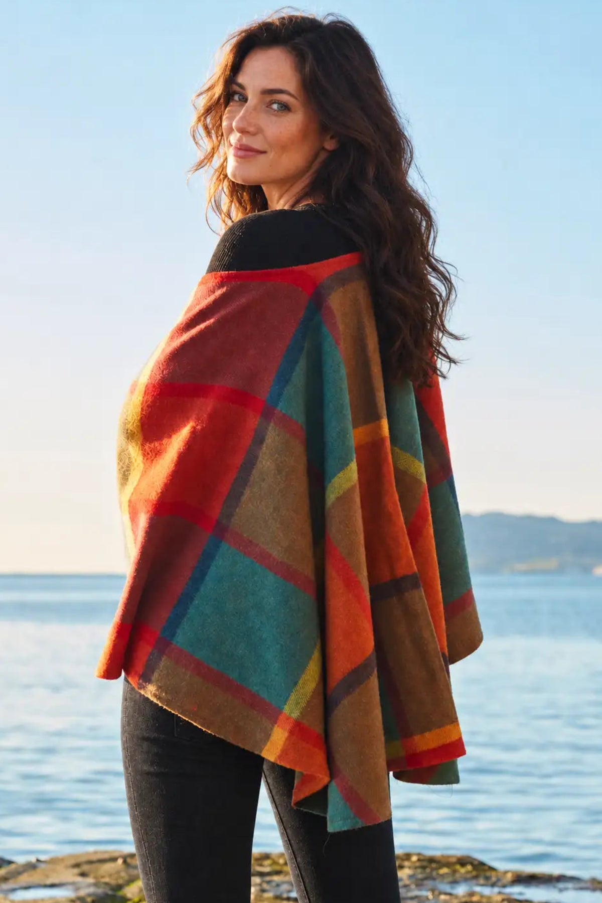 A woman with long brunette wavy hair wearing a plaid red, blue, teal and yellow Irish wrap by the coast.