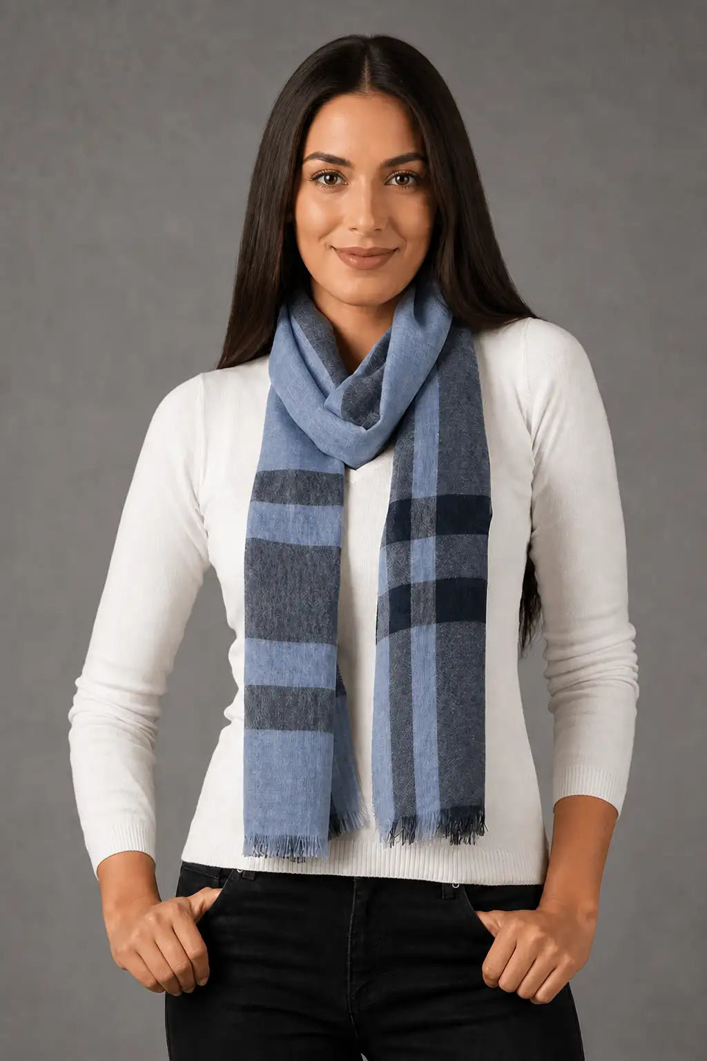 A woman modeling a light blue and navy plaid scarf wrapped around her neck, paired with a white long-sleeve top and black pants against a gray studio background.
