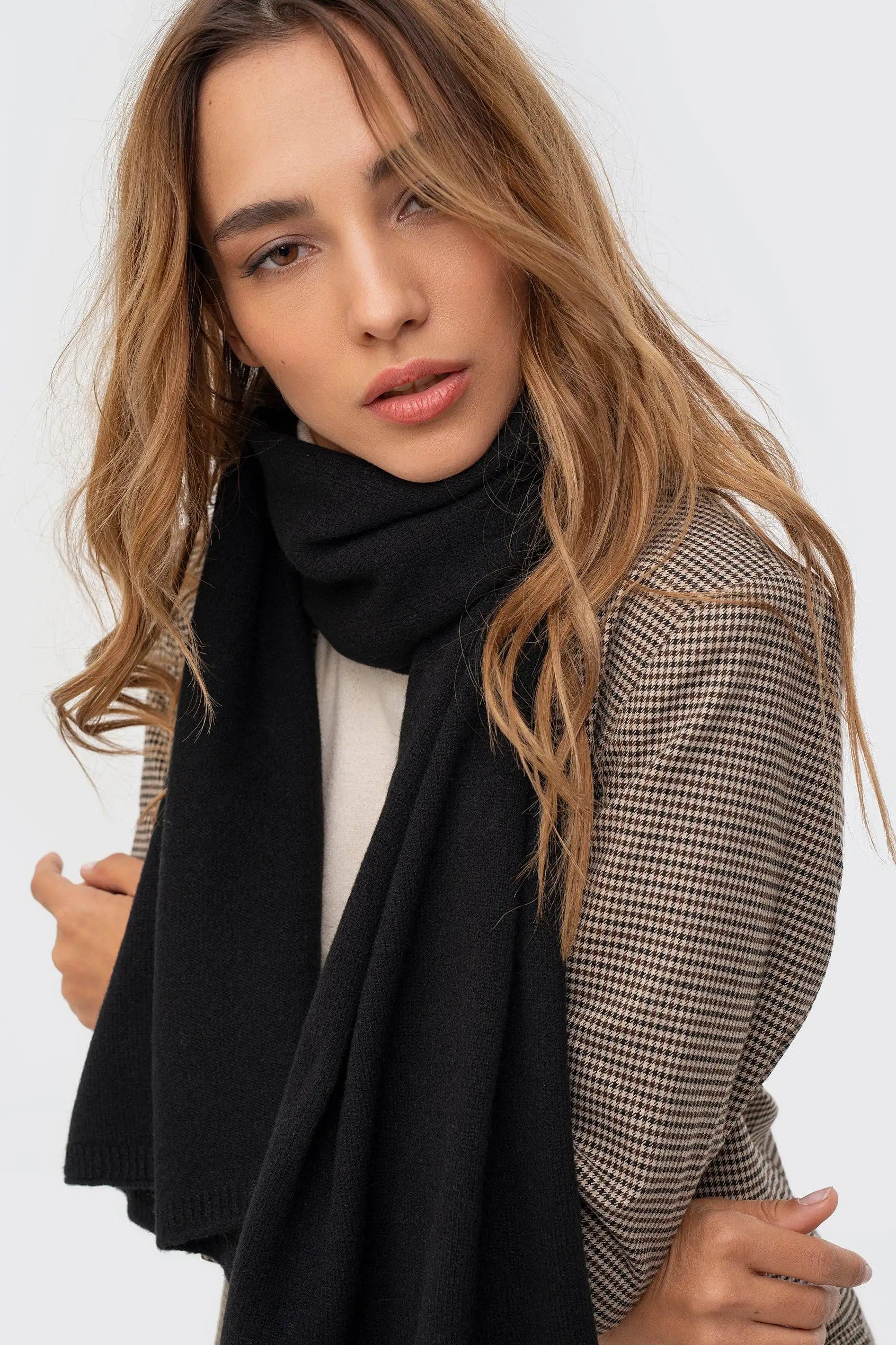 A woman wearing a thick black knit scarf and a brown and beige houndstooth blazer, photographed against a clean light gray background.