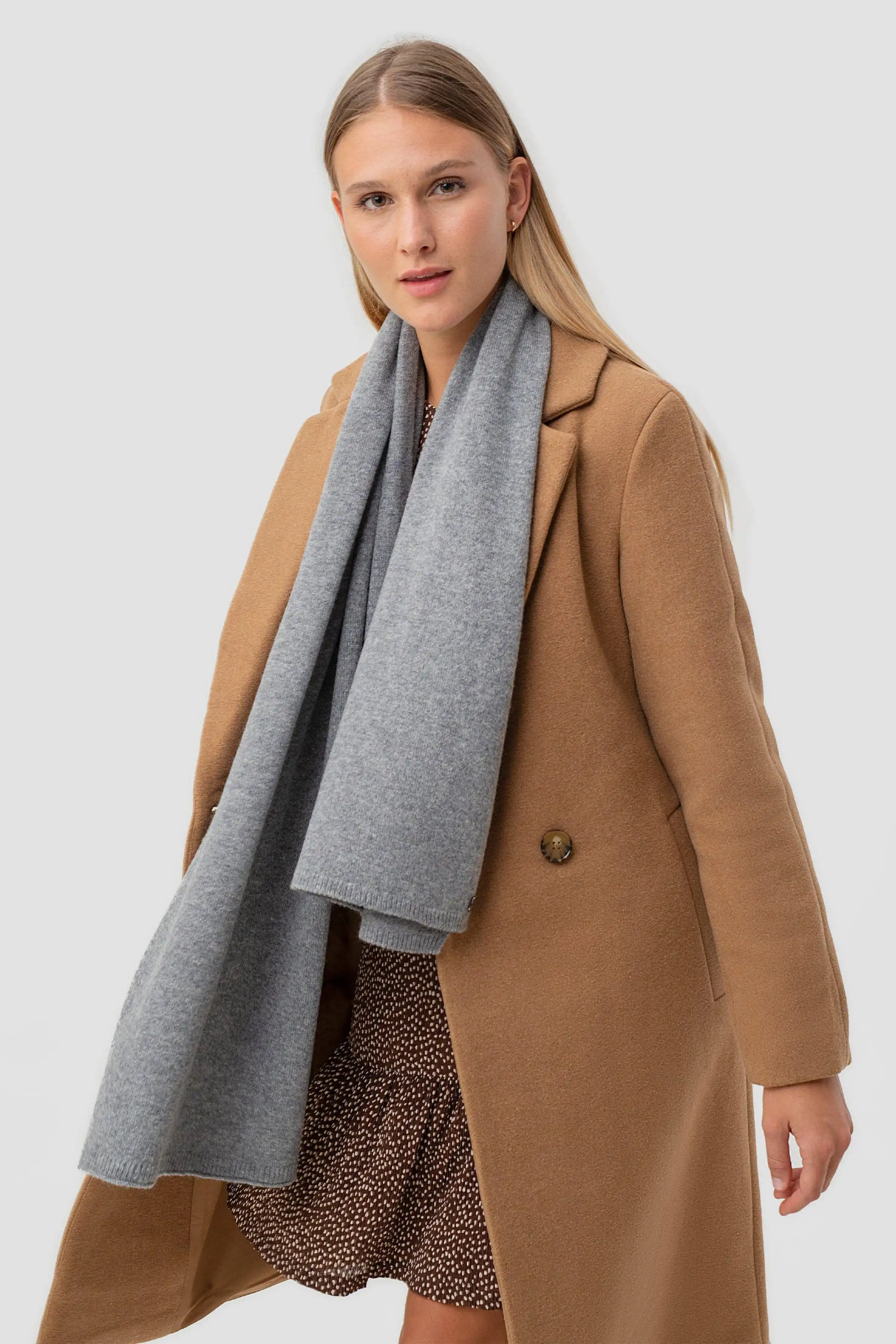 A woman modeling a long camel-colored coat with a wide, soft gray scarf wrapped around her neck, set against a solid light gray studio background.