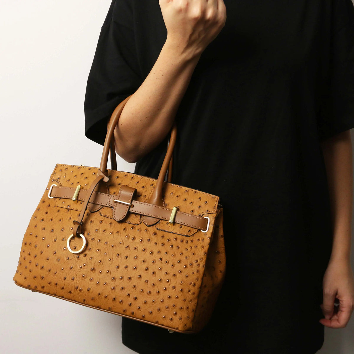 Acognac ostrich-textured leather handbag with gold-tone hardware, carried on the arm of a person wearing a black t-shirt against a plain white background.