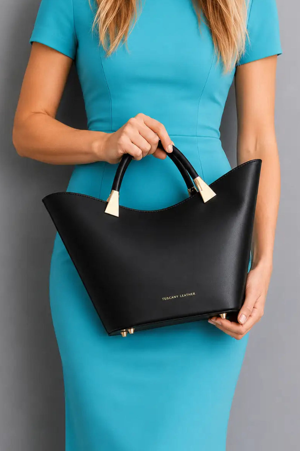 A woman in a bright turquoise dress holds a structured black leather handbag with double top handles and small gold-tone hardware against a plain gray studio background.