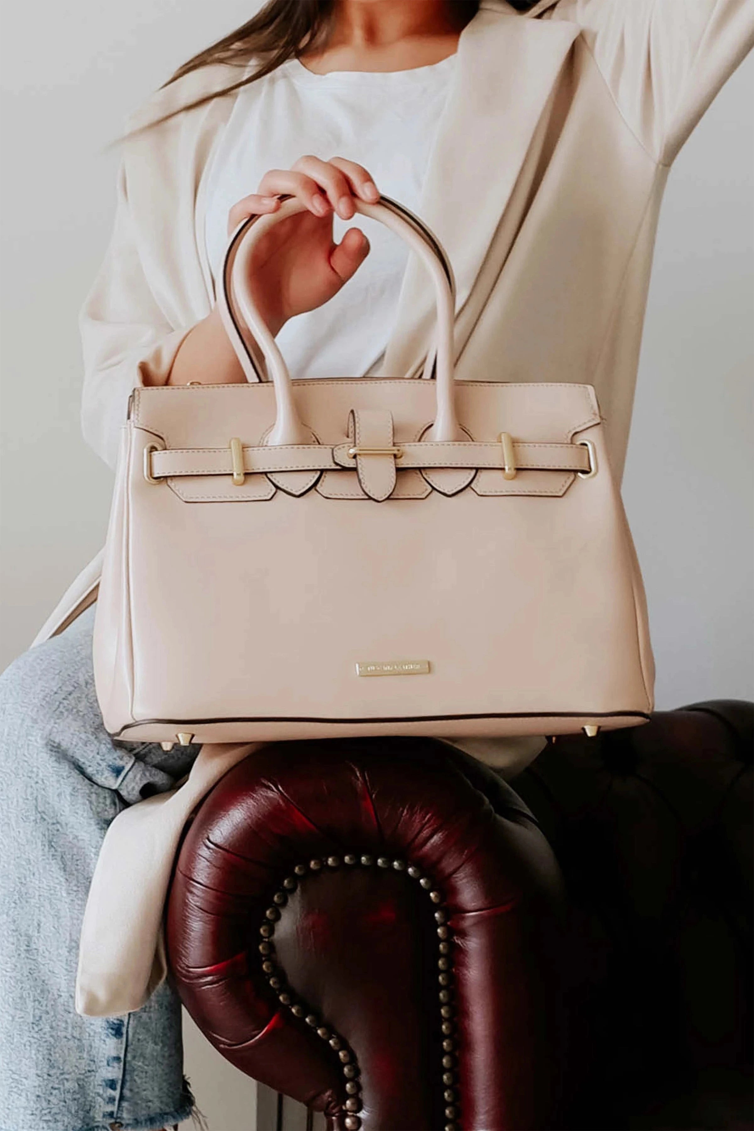 A woman is sitting on a brown leather chair wearing a cream sweater and blue jeans holding a Cream leather bag.