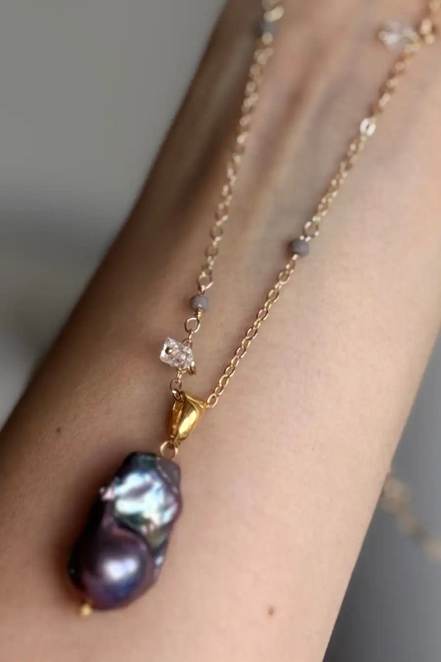 A gold chain necklace with a baroque pearl pendant and small clear stones, shown worn on a person's wrist.