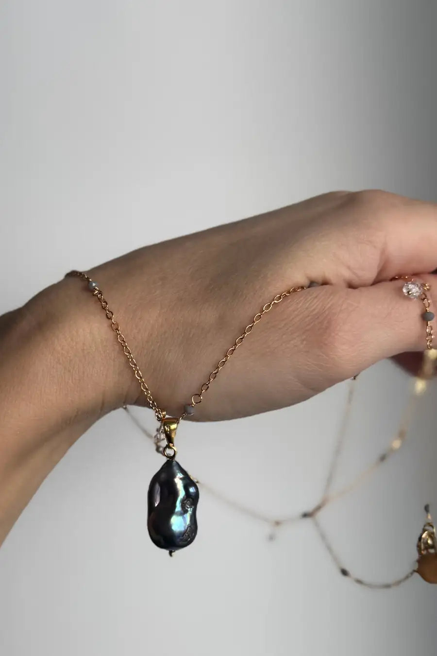 A gold chain necklace with a large, irregular Tahitian baroque pearl pendant featuring iridescent blue-green flashes, held on a person's wrist against a neutral light gray background.