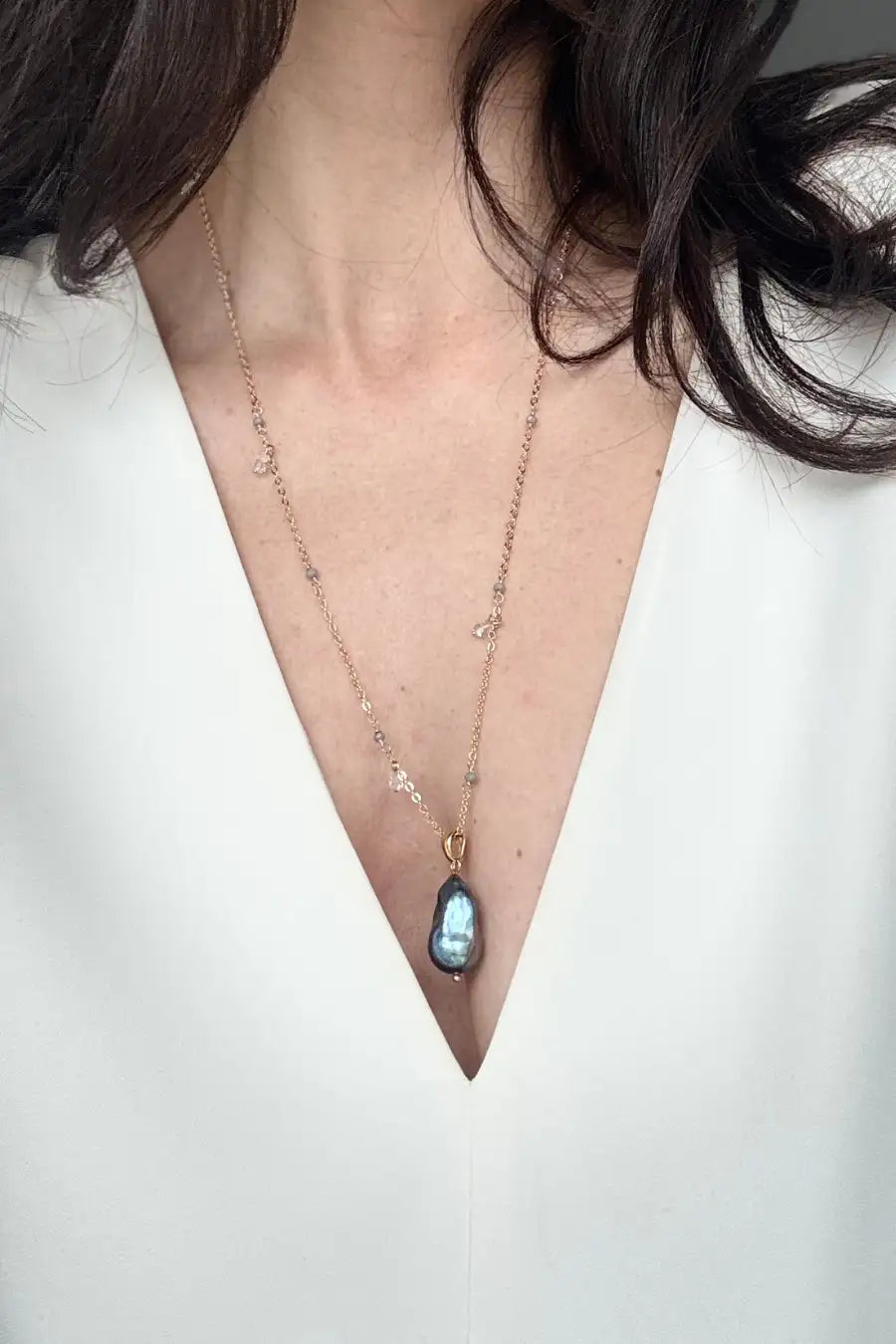 A close-up of a model wearing a delicate gold chain necklace with a teardrop-shaped iridescent blue pearl pendant, styled with a white V-neck top.