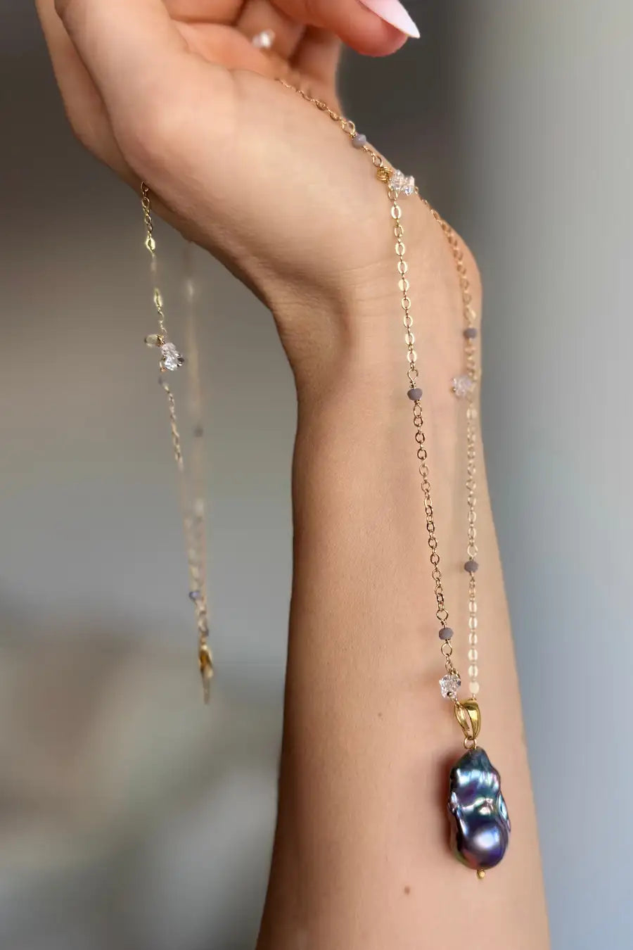A delicate gold chain necklace with a large, irregular dark blue-black baroque pearl pendant, held by a person's hand against a soft, blurred gray background.