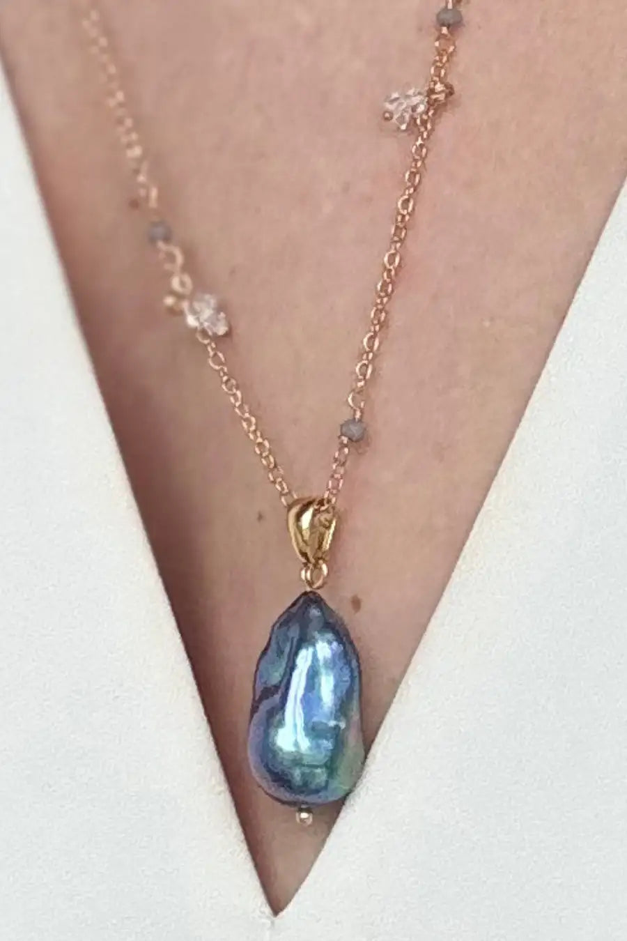 A close-up of a delicate necklace worn on a person's neck, featuring a teardrop-shaped iridescent blue-green pearl pendant with a gold-tone setting and a rose gold chain accented with small clear crystals.