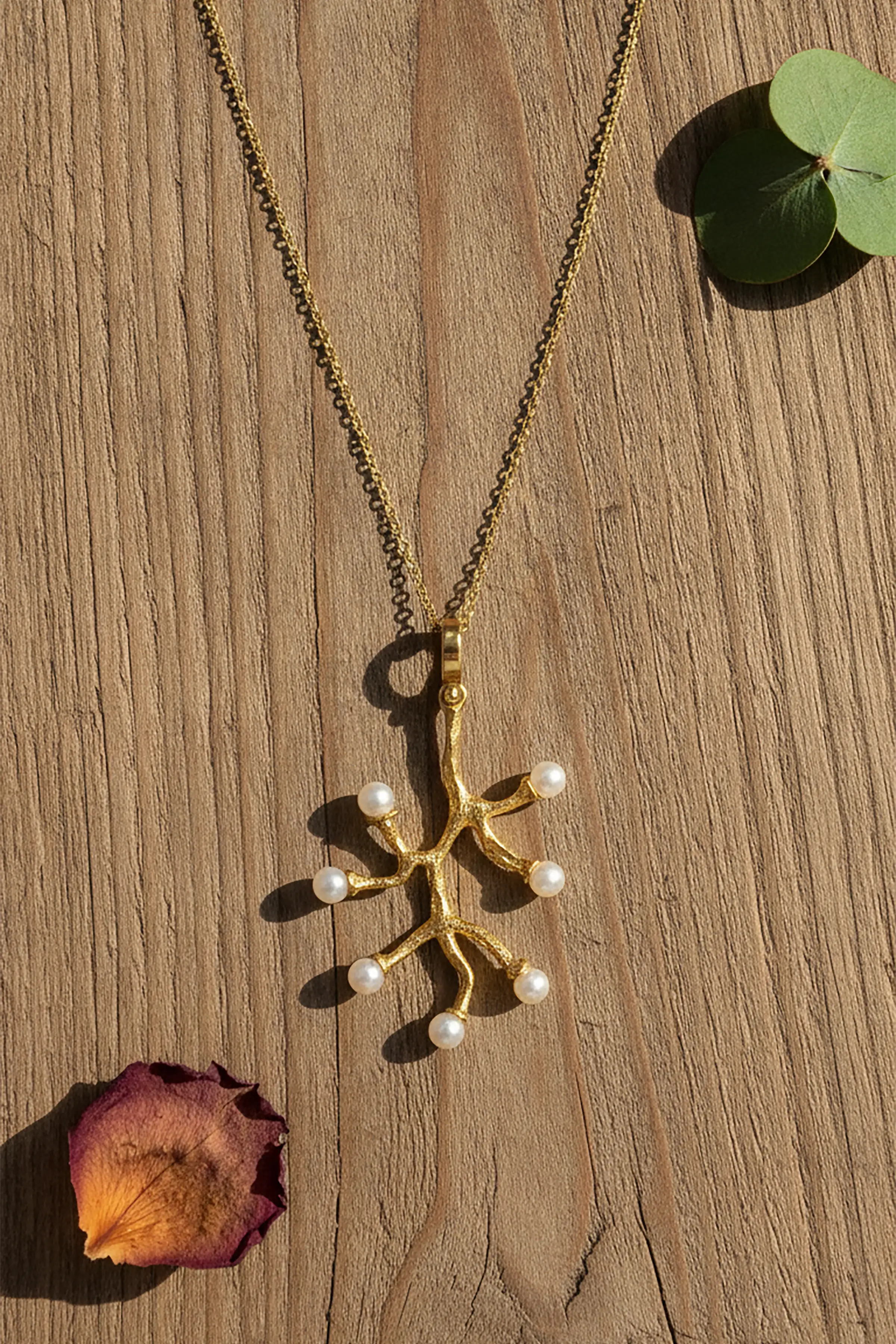 A gold branch-shaped pendant necklace with small white pearl accents, displayed on a wooden surface with decorative green leaves and a dried flower petal.