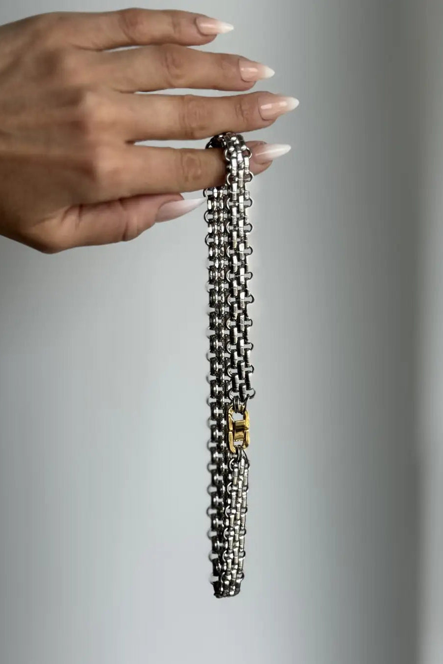 A close-up of a hand holding a silver-toned chain necklace with a small gold accent clasp against a neutral gray background.