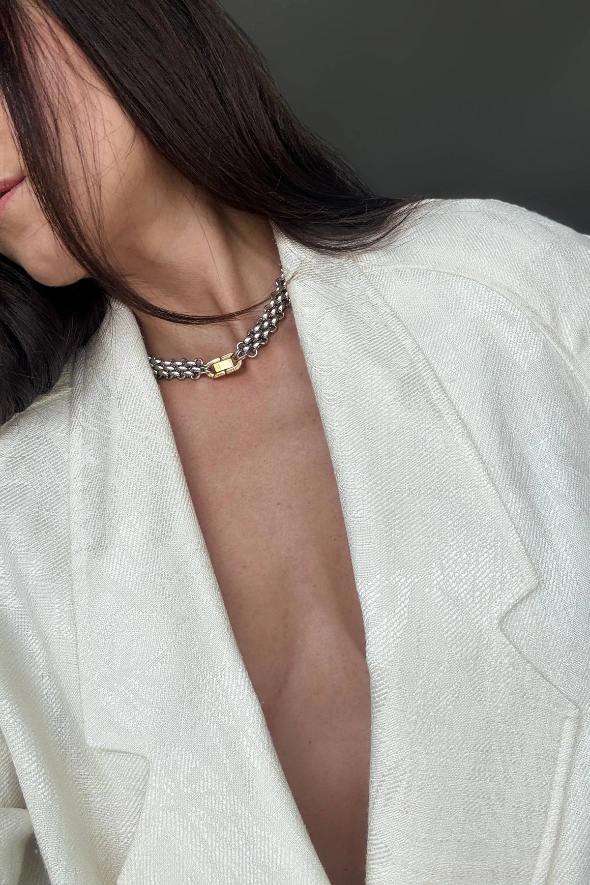 A close-up of a model wearing a silver chain choker necklace with a small gold heart charm, styled with a textured off-white blazer.