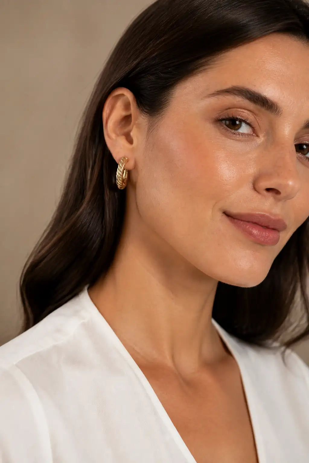 A close-up of a woman wearing a small textured gold hoop earring, dressed in a white V-neck top against a soft beige background.