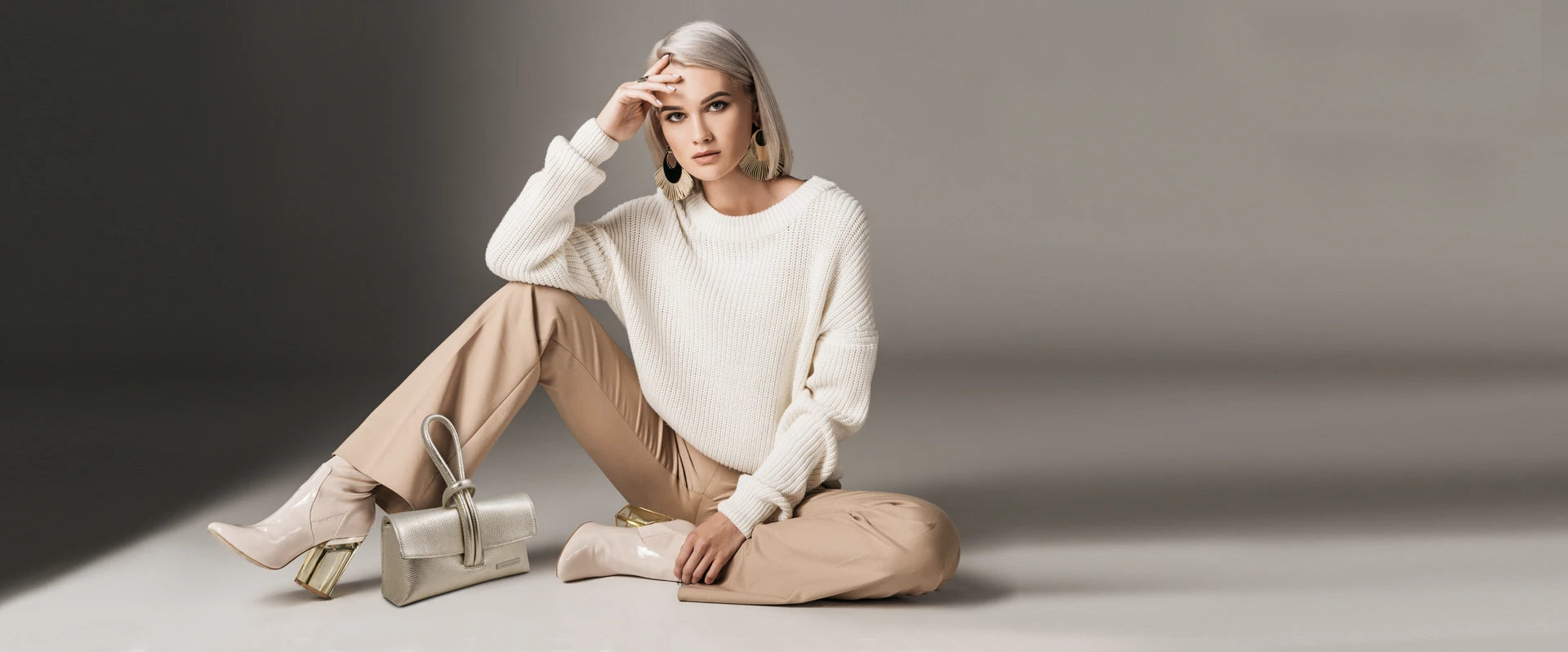 A woman wearing a cream sweater and beige pants sitting on floor next to a silver clutch.