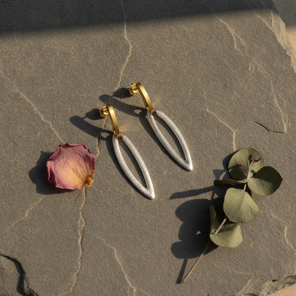 A pair of gold hoop earrings with textured silver teardrop dangles, styled on a textured stone surface with a dried rose petal and green leaves.