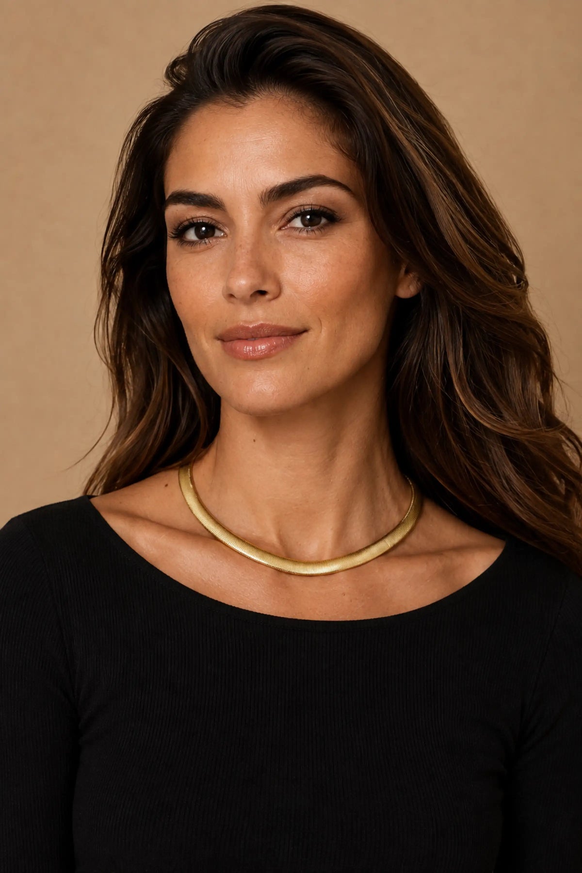 A woman with long brown hair wearing a gold 16" snake chain necklace and a black top, photographed against a warm beige background.