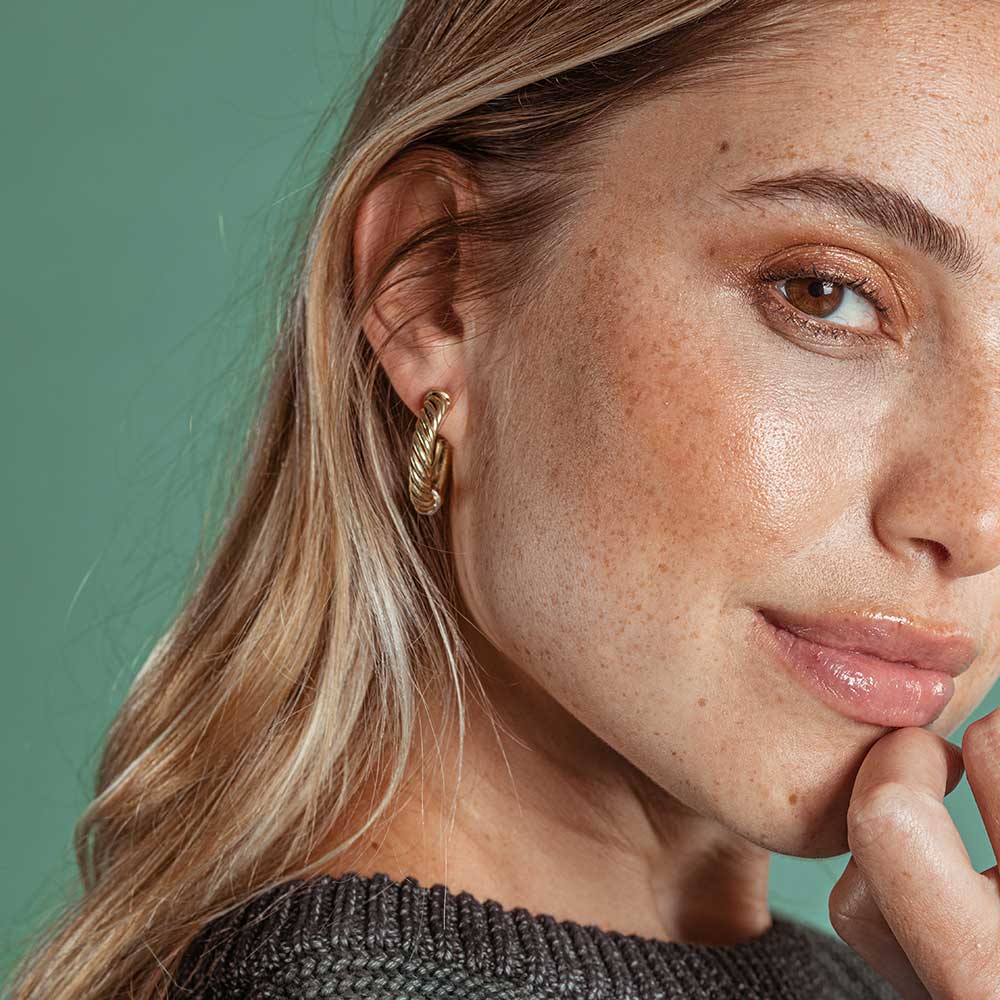 A close-up of a woman with blonde hair wearing a textured gold hoop earring, set against a solid teal-green background.