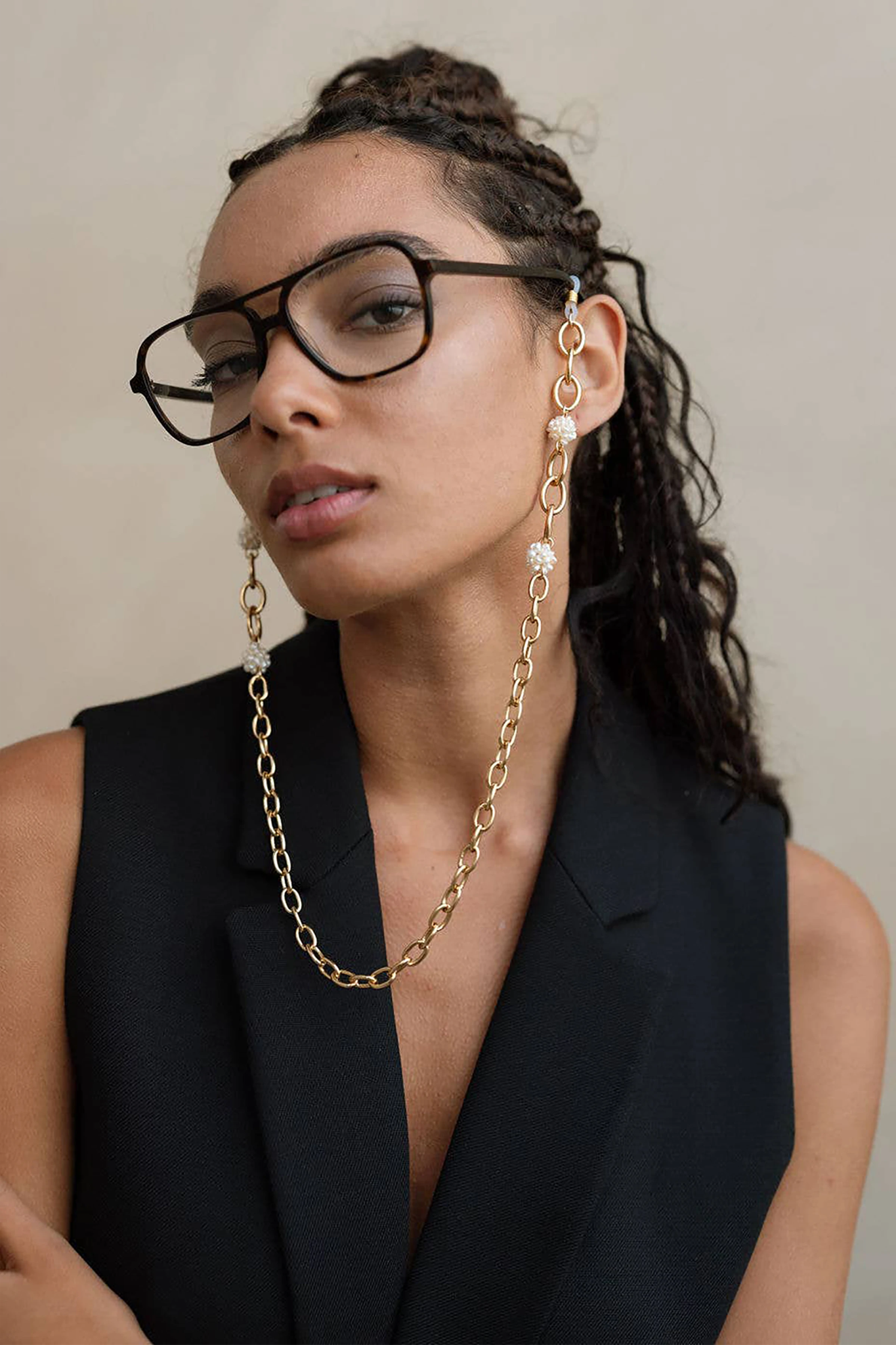 Esmee Golden Pearl Eyeglass Chain on a model wearing a black sleeveless suiting vest and black large-rimmed glasses. The chain is in front of her.