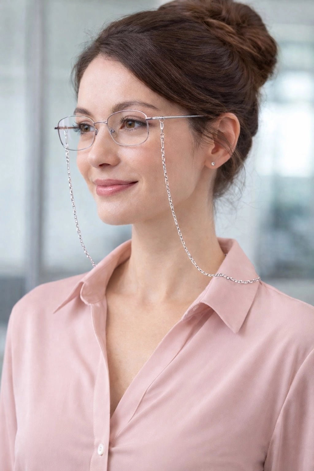 Woman wearing a light pink shirt, glasses with a silver chain, standing indoors in front of office windows.