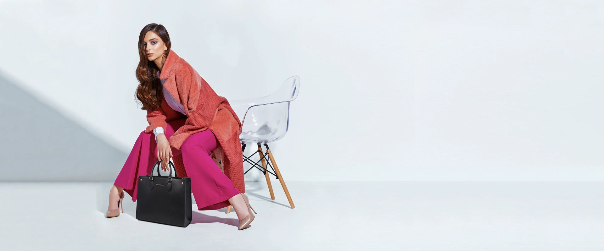 Woman wearing orange long jacket, fuchsia pants, nude heels holding black briefcase on light gray background